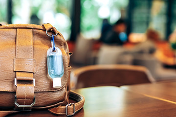 Mini portable alcohol gel bottle to kill Corona Virus(Covid-19) hang on a brown leather shoulder bag on table in coffee shop.New normal lifestyle. Health care concept. Selective focus on alcohol gel