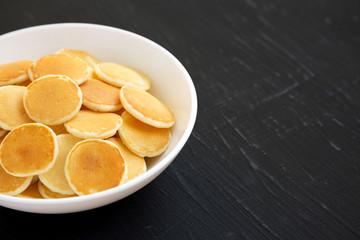 Homemade Mini Pancakes Cereal in a white bowl on a black background, side view. Copy space.