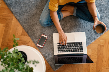 woman in a yellow shirt works on a laptop with a mug of coffee at home in the living room on the carpet, remote work and education, top view