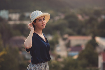 Portrait of elegant woman holds her hat in her hand and poses in a half-turn. Side view. In the background, a view of the city. Copy space. Travel and leisure concept