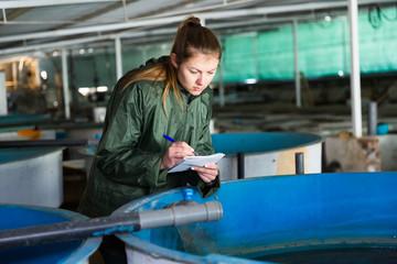 Woman owner of trout farm checking reservoirs with fry, writing results of inspection in notebook..