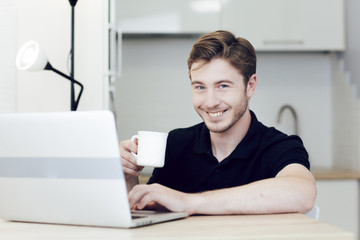Smiling young man working at home on laptop sitting at desk and holding a cup of coffee. Work at a distance during quarantine of coronavirus. Stay at home, self-isolation