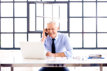 Naklejka na ściany i meble Elderly businessman talking with somebody on his mobile phone while working on laptop