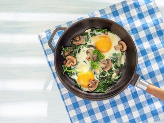 Cooking pan with spinach, mushrooms, cheese and fried eggs. Healthy homemade dish for low carb diet on a light blue wooden table with linen towel. Close up, flat lay. Top view. Idea for breakfast