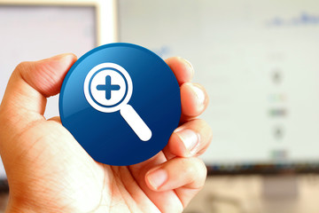 Zoom in icon blue round button holding by hand infront of workspace background