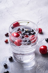 Fresh cold sparkling water drink with cherry, raspberry and currant berries in transparent glass on stone concrete background, summer diet beverage, angle view macro