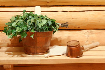 A fresh birch broom and copper vat and ladle are on a wooden bench in sauna.