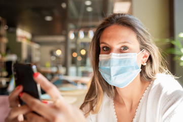 Young blonde Caucasian girl in a face mask sitting at a bar, looking at the camera while holding her smart phone in her hands. Copy space.Pandemic and technology concept