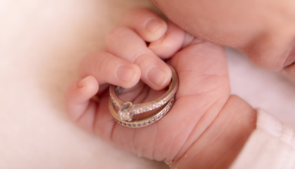 Newborn baby little hand holds parents' gold and silver wedding rings on its palm on white blanket photography.