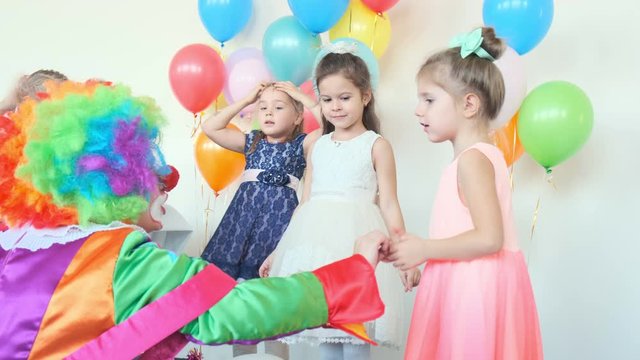 funny clown in rainbow wig entertains cute little girls standing near wall with air balloons at birthday party slow motion