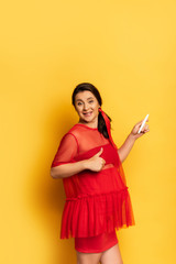 young pregnant woman in red tunic showing thumb up while holding pregnancy test on yellow
