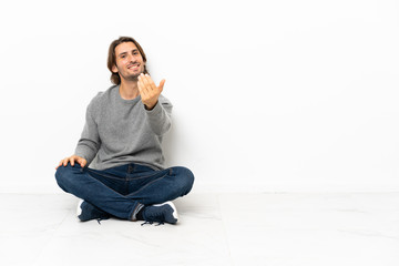 Young handsome man sitting on the floor over isolated background inviting to come with hand. Happy that you came