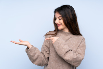 Young brunette woman wearing a sweater over isolated blue background holding copyspace imaginary on...