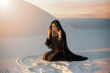 A mysterious woman in a black long dress sits in the desert. Luxurious clothes, gold accessories...