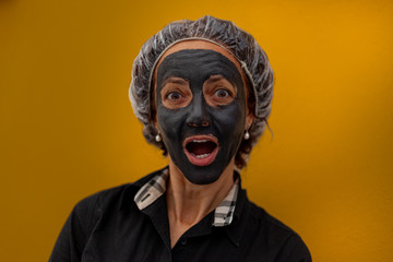 Brazilian woman with surprised expression and gray clay mask. Concept: beauty, face care, facelift, wellness, skin care. Shot with orange background
