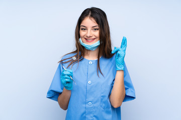 Woman dentist holding tools over isolated blue background with fingers crossing