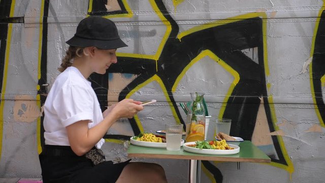 Side view of a cute girl in a bucket hat and white t-shirt taking a picture of her meal sitting next to graffiti wall in urban cafe. 