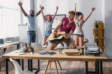 Selective focus of business people jumping while colleague playing acoustic guitar in office