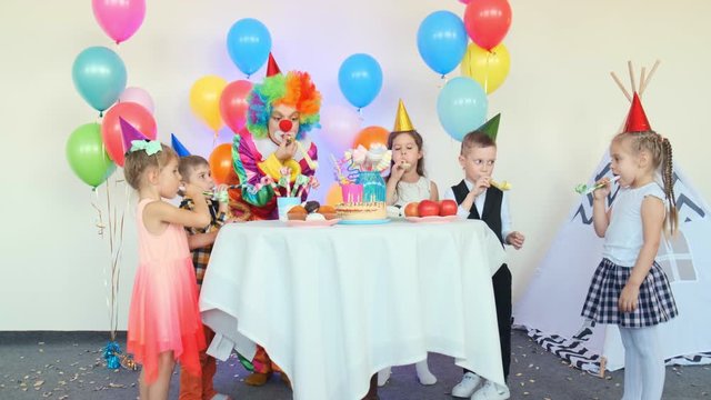 positive little children with funny clown blow party horns at table with holiday cake in room at birthday party slow motion