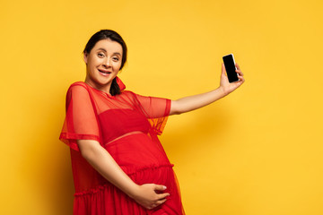 brunette pregnant woman in red tunic taking selfie on smartphone while looking at camera on yellow