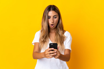 Young hispanic woman over isolated yellow background looking at the camera while using the mobile with surprised expression