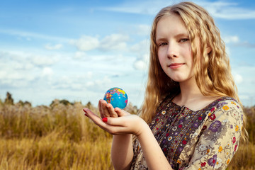 A  girl with curly wheat hairs  holds  toy of the globe , face close-up, on yellow field and blue sky. Travel concept. 