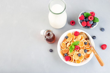 Small cereal pancakes with maple syrup, raspberry, blueberry and milk, in white bowl, horizontal, top view, copy space