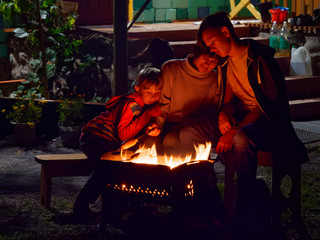 Group of young friends sitting by the fire late at night, grilling sausages and having fun