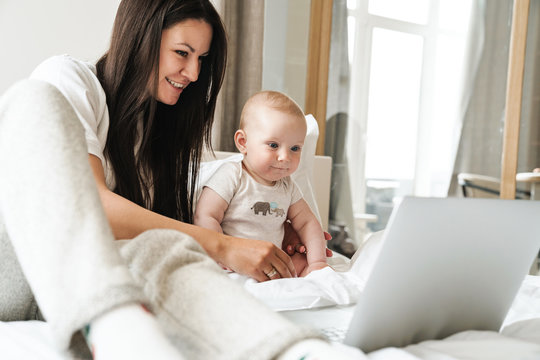 Photo of happy mother using laptop and playing with her baby on bed