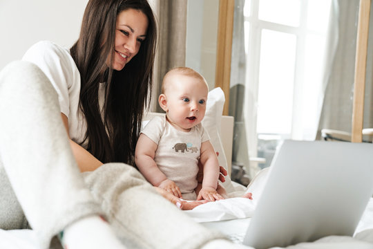 Photo of happy mother using laptop and playing with her baby on bed