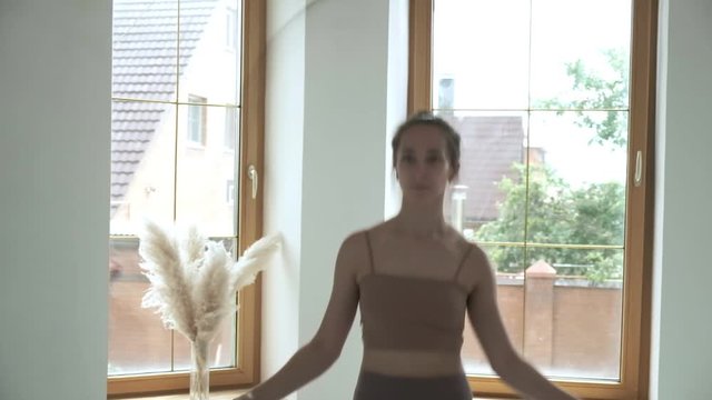 Young fit brunette girl with ponytail in beige top, leggings does jump exercise in bright room with skipping rope. sports, fitness training at home. Healthy lifestyle. slim woman in sportswear jumping