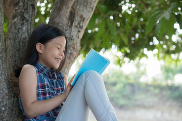 little asian girl reading a book under big tree. children and science. blurred background. learning the imagination and dreams of rural child. studying at home.