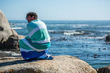 young man sitting on the beach wrapped in a towel