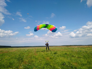 A skydiver with a bright multicolored parachute flies against the background of a blue sky with white sparse clouds and green grass in summer