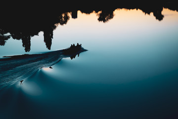 Silhouette of a motor, rubber boat at sunset over the lake. Smooth water surface torn by the waves. Water reflection of fishermans pontoon and forest on the far shore.