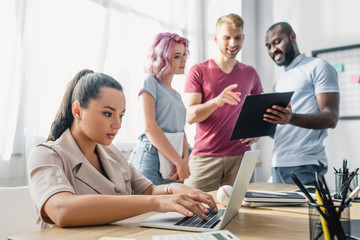 Selective focus of businesswoman using laptop while colleagues working with clipboard in office