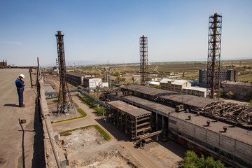 Fototapeta na wymiar Old unused soviet metallurgy factory buildings and rusted chimneys on the blue sky. Worker in white hardhat (left) on the roof. Taraz city, Kazakhstan.