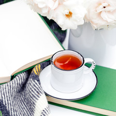 Cup of tea on books, flowers white wild rose in vase , warm plaid on white table outside in garden. Romantic leisure breakfast with nature background. Cozy home backyard or provence cafe