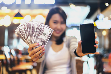 Young woman showing her mobile phone with a bunch of dollar banknotes. Make money online with...