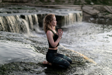 Blonde young woman in sportswear practicing breathing yoga outdoors in harmony with nature. Fitness girl meditating with closed eyes on the edge of waterfall with hands clasped in namaste mudra sign.