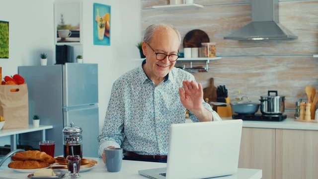 Senior man chatting and waving during a video call using laptop in kitchen holding a cup of coffee. Elderly person using internet online chat technology video webcam making a video call connection