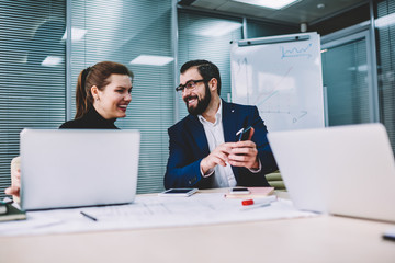 Young handsome smiling graphic designer with attractive female colleague watching funny video in web page on smartphone.Cheerful coworkers sincerely laughing while reading news in website on cellular