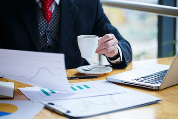 Adult male businessman working on a new project and looking at stock growth charts. Sits at a large window at the table. Looks at the laptop screen and drinks coffee.
