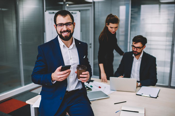 Portrait of beautiful attractive young business man in eyeglasses with beard is in office with colleagues on background, prosperous man sitting on table drink coffee smiling and looking at camera