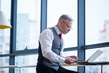 Adult male businessman, teacher, mentor working on a new project. Sits by a large window on the table. He looks at the laptop screen.