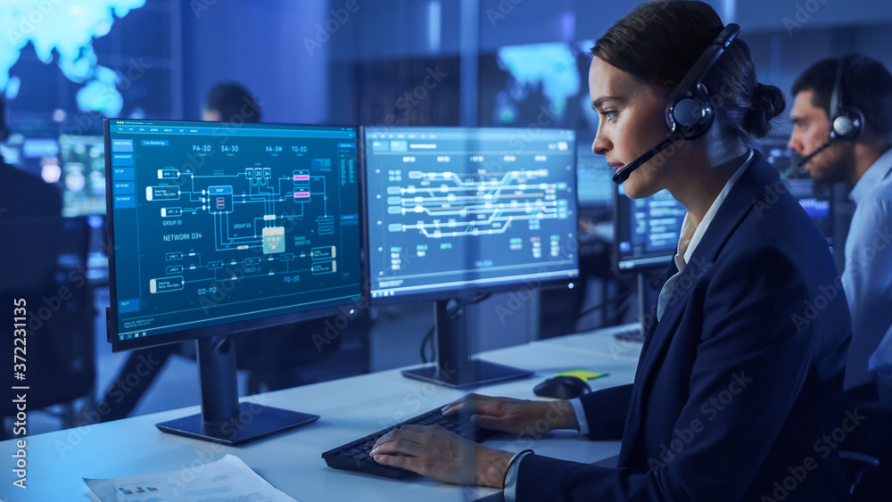 Poster Confident Female Data Scientist Works on Personal Computer Wearing a Headset in Big Infrastructure Control and Monitoring Room. Woman Engineer Works with Network Logics in Office Room with Colleagues.