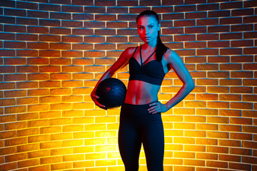 Serious sporty young brunette woman posing with medicine ball in gym in neon lights.