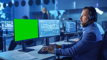 Confident Male Data Scientist Works on Computer with Green Screen Mock Up Template in Big Infrastructure Control and Monitoring Room. Senior Engineer in Call Center Office Room with Colleagues.