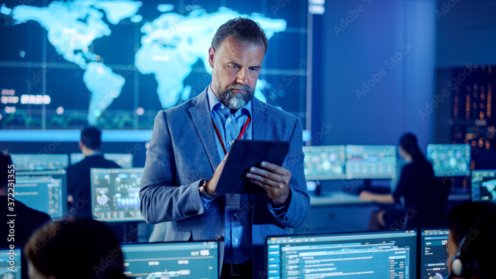 Aufkleber Male Project Leader is Standing with Tablet Computer and Checking Work Data. Science Engineers Work Around Him. Telecommunications Control Monitoring Room with a Global Map on Big Screen.