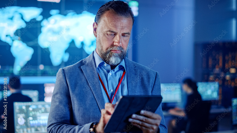 Poster Male Project Leader is Standing with Tablet Computer and Checking Work Data. Science Engineers Work Around Him. Telecommunications Control Monitoring Room with a Global Map on Big Screen.